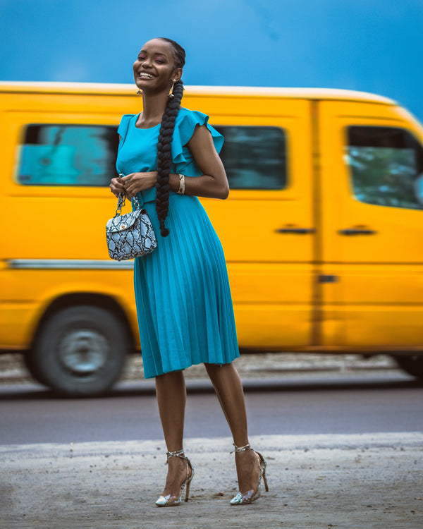 A smiling black African woman in a blue pleated mide dress clutching a cute handbag